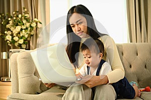Asian mother holding her baby boy and working with laptop on couch at home