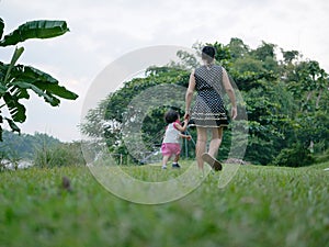 Asian mother holding her little daughter`s hand and have a walk together on an outdoor green grass field by riverside