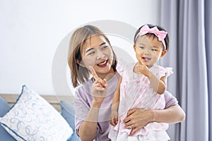 Asian mother is holding her pretty smiling baby daughter while spending quality time in the bed for family happiness and parenting