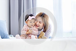 Asian mother is holding her pretty smiling baby daughter while spending quality time in the bed for family happiness and parenting