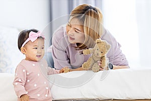 Asian mother is holding her pretty smiling baby daughter while spending quality time in the bed for family happiness and parenting