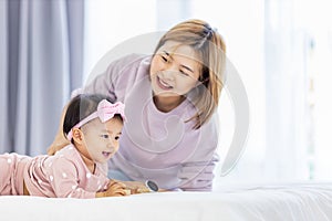 Asian mother is holding her pretty smiling baby daughter while spending quality time in the bed for family happiness and parenting