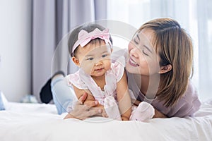 Asian mother is holding her pretty smiling baby daughter while spending quality time in bed for family happiness and parenting