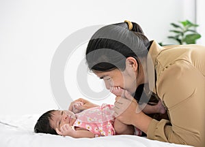 Asian mother holding and kissing newborn baby foot with love. Infant girl lying on bed at home and holding mom finger. Love and