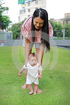 Asian mother learning her infant baby to walk first steps on the green grass garden