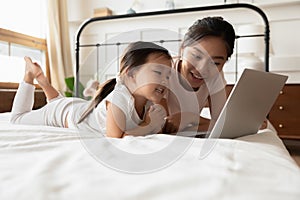 Asian mother and little daughter lying in bed with laptop