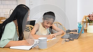 Asian mother and little daughter sitting in kitchen studying online, learning internet online class and doing homework