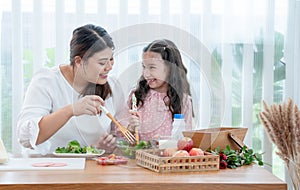 Asian mother and little helper, Caucasian child daughter preparing breakfast in kitchen, mixing vegetables and fruit in salad bowl