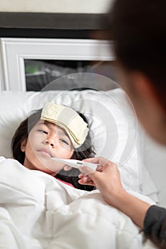 Asian mother measuring temperature girl with digital thermometer on bed at morning time, Sick child have cool towel for reduce