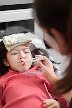 Asian mother measuring temperature girl with digital thermometer in her mouth on bed at morning time, Sick child have cool towel