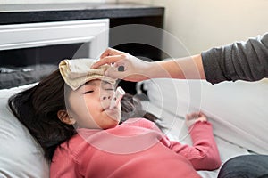 Asian mother measuring temperature girl with digital thermometer in her mouth on bed at morning time, Sick child have cool towel