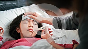 Asian mother measuring temperature girl with digital thermometer in her mouth on bed at morning time, Sick child have cool towel