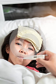 Asian mother measuring temperature girl with digital thermometer in her mouth on bed at morning time, Sick child have cool towel