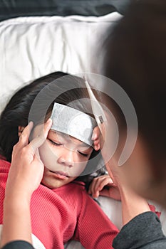 Asian mother measuring temperature girl with digital thermometer in her mouth on bed at morning time, Sick child have cool towel