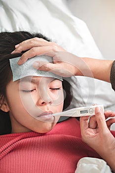 Asian mother measuring temperature girl with digital thermometer in her mouth on bed at morning time, Sick child have cool towel