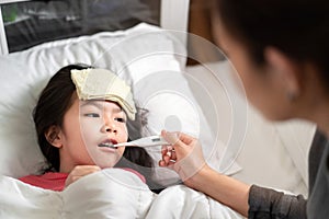 Asian mother measuring temperature girl with digital thermometer in her mouth on bed at morning time, Sick child have cool towel