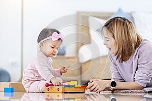 Asian mother is playing with her pretty smiling baby daughter with wooden toy block while spending quality time in the bed for