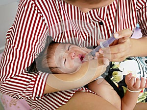 Asian mother`s arm wrapping around her crying baby girl`s face forcing the baby to take liquid medicine