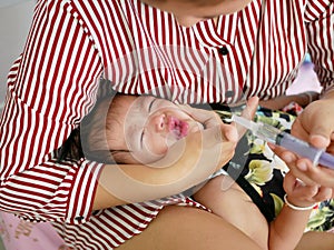 Asian mother`s arm wrapping around her crying baby girl`s face forcing the baby to take liquid medicine