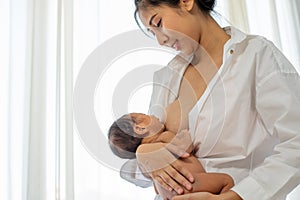 Asian mother with smiling give breastfeeding newborn with white shirt and sitting on bed white background of glass windows and