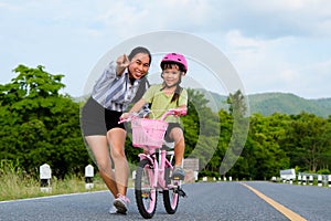 Asian mother teaching daughter to ride a bicycle in the park. A cute happy little girl learning to ride a bicycle with her mother