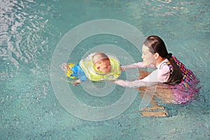 Asian Mother training for infant baby boy in swimming suit floating in pool with safety by baby neck floats