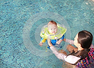 Asian Mother training for infant baby boy in swimming suit floating in pool with safety by baby neck floats