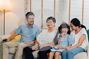 Asian multi generational family of father, mother, daughter, and grandmother watching TV on sofa in the living room