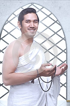 Asian Muslim man in ihram clothes standing and praying with prayer beads while raised arms