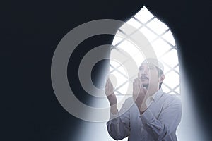 Asian Muslim man sitting while raised hands and praying inside the room