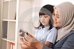 Asian muslim mom and little baby girl daughter learning online or watching videos on tablet phone, happiness between mother and