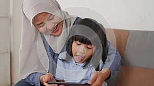 Asian muslim mom and little baby girl daughter learning online or watching videos on tablet phone, happiness between mother and