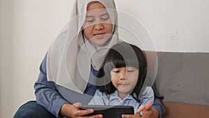 Asian muslim mom and little baby girl daughter learning online or watching videos on tablet phone, happiness between mother and