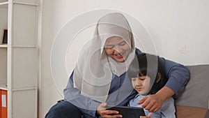 Asian muslim mom and little baby girl daughter learning online or watching videos on tablet phone, happiness between mother and