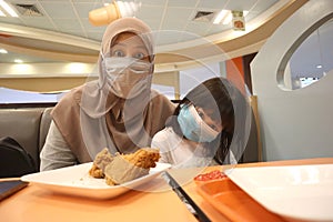 Asian muslim mother and baby girl daughter wearing protective face mask at fast food restaurant. Eating dine in during coronavirus