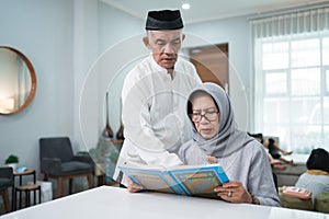 Asian Muslim senior man teaching wife reading Koran or Quran in living room