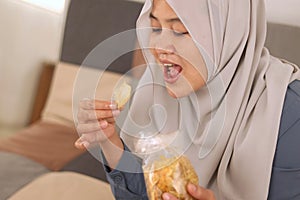 Asian muslim woman enjoys eating casava or potato chips while sitting on sofa at living room