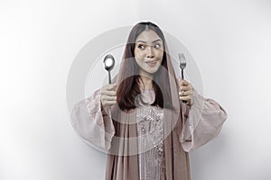 An Asian Muslim woman is fasting and hungry and holding cutlery while looking aside thinking about what to eat