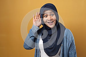 Asian muslim woman wearing hijab smiling while doing hearing gesture, shocked surprised expression to hear gossip