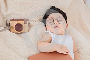 Asian Newborn baby wearing glasses with favorite book on beige blanket , 3 month-old infant lying in bed with relax. Adorable baby