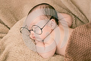 Asian Newborn baby wearing glasses and sleep on brown bed at home, 0-1month-old infant lying in blanket with relax. Adorable baby