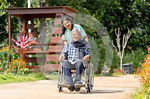 Asian nurse cart of shove senior man sit on wheelchair to walk along the walkway in the garden and they look happiness outdoor to