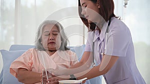 Asian nurse helping elderly woman patient to drink on bed at hospital ward