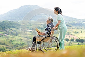 Asian nurse stand behide and hold wheelchair handle with senior man sit on wheelchair and both look forward and stay near cliff