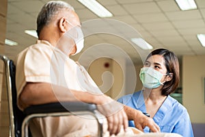 Asian nurse taking care of mature male patient sitting on wheelchair in hospital. Young woman and old man wearing surgical face