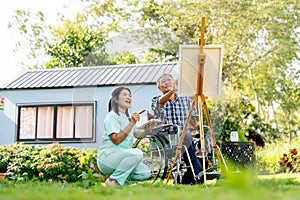 Asian nurse woman sit near senior man on wheelchair and cheer up him to paint the painting outdoor in the garden for relax and get