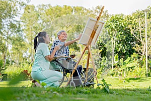 Asian nurse woman sit near senior man on wheelchair enjoy to paint the painting outdoor in the garden and they look happiness