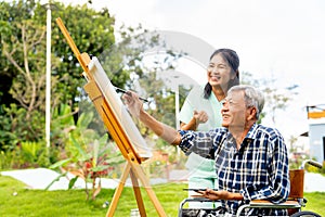Asian nurse woman stand in the back to cheer up senior man on wheelchair to paint the painting outdoor and they look happy
