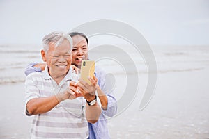 Asian old senior couples use smartphone to selfie at the beach by the sea background on weekend vacation.Concept of happy living