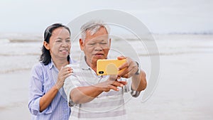 Asian old senior couples use smartphone to selfie at the beach by the sea background on weekend vacation.Concept of happy living